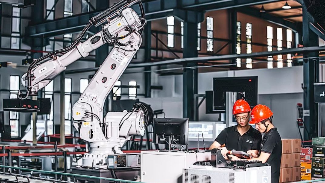 An automated industrial robot next to two workers controlling it with a laptop
